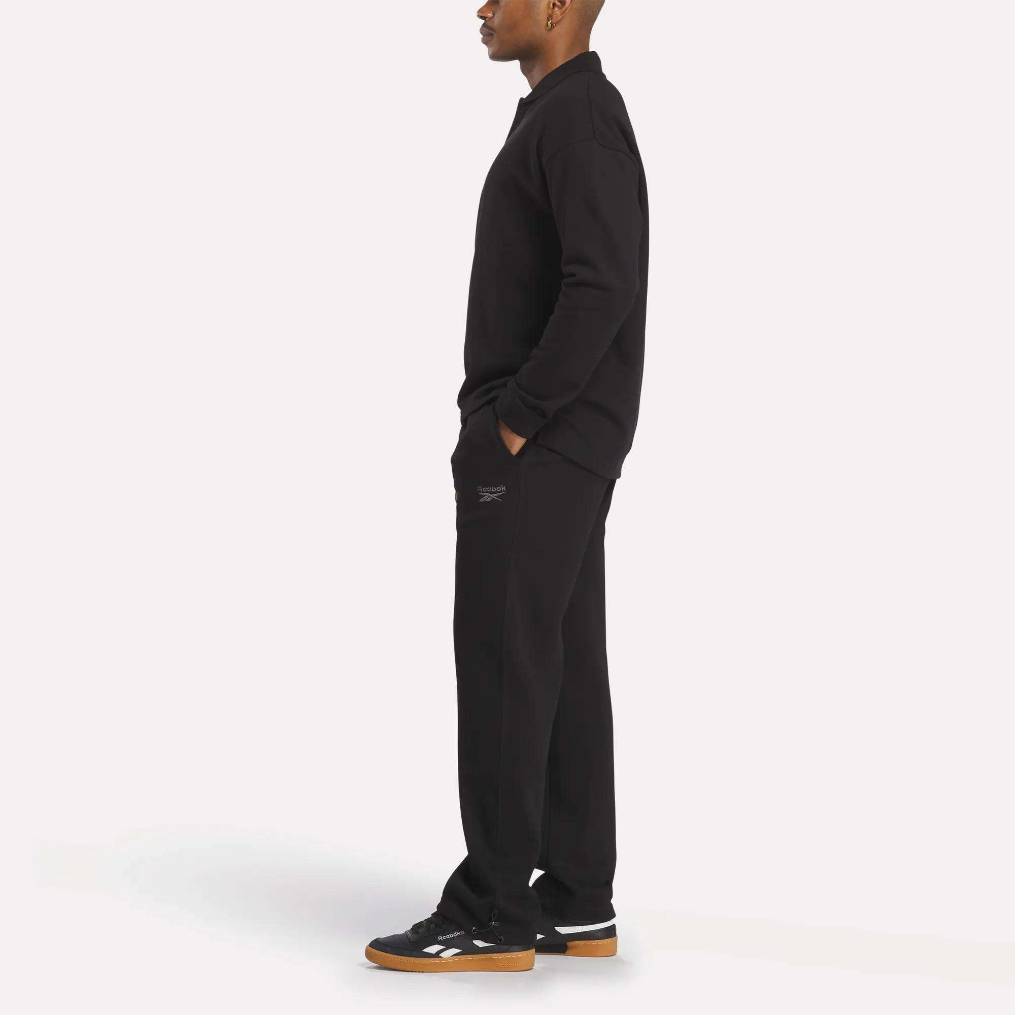 A person in profile against a white background wears a relaxed fit Men’s Wardrobe Essentials Long Sleeve Polo Shirt by Reebok, paired with black pants (hands in pockets) and black sneakers featuring white details and gum soles.