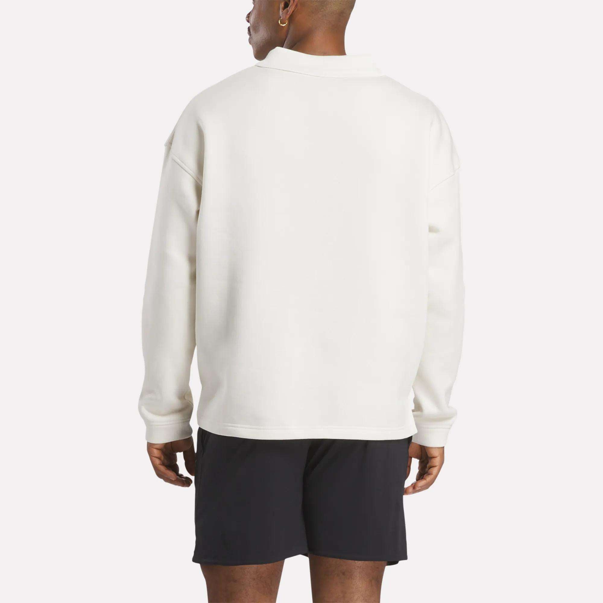 A person stands with their back to the camera, wearing a Reebok Men's Wardrobe Essentials Long Sleeve Polo Shirt in white and black shorts against a plain white background.