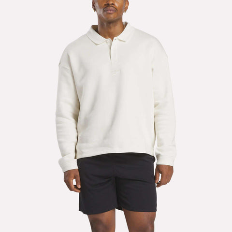 A person wearing a Reebok Men's Wardrobe Essentials Long Sleeve Polo Shirt and black shorts stands against a plain white background.