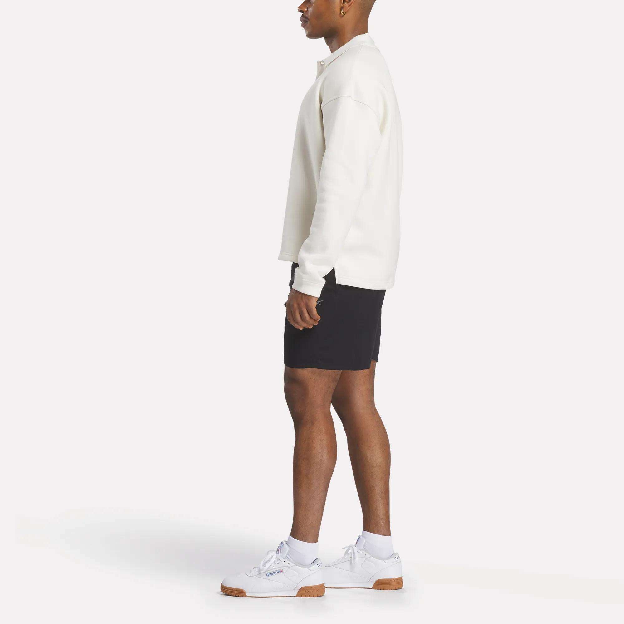 A man in profile wears the Reebok Men's Wardrobe Essentials Long Sleeve Polo Shirt, paired with black shorts and white sneakers, set against a plain white background.
