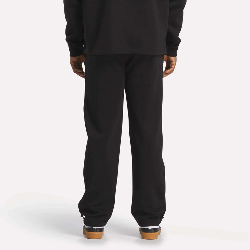 A person faces away, showing only their lower body in Reebok Men's Wardrobe Essentials Pants, black cotton-blend fleece. They're paired with black and white sneakers with gum soles against a plain white background.