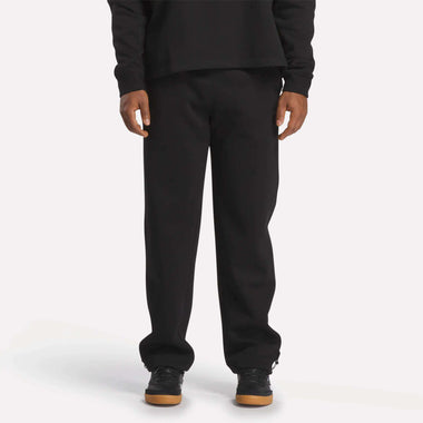 A person wearing Reebok Men's Wardrobe Essentials Pants in black, a matching black long-sleeve shirt, and black sneakers with light brown soles, standing against a plain white background.