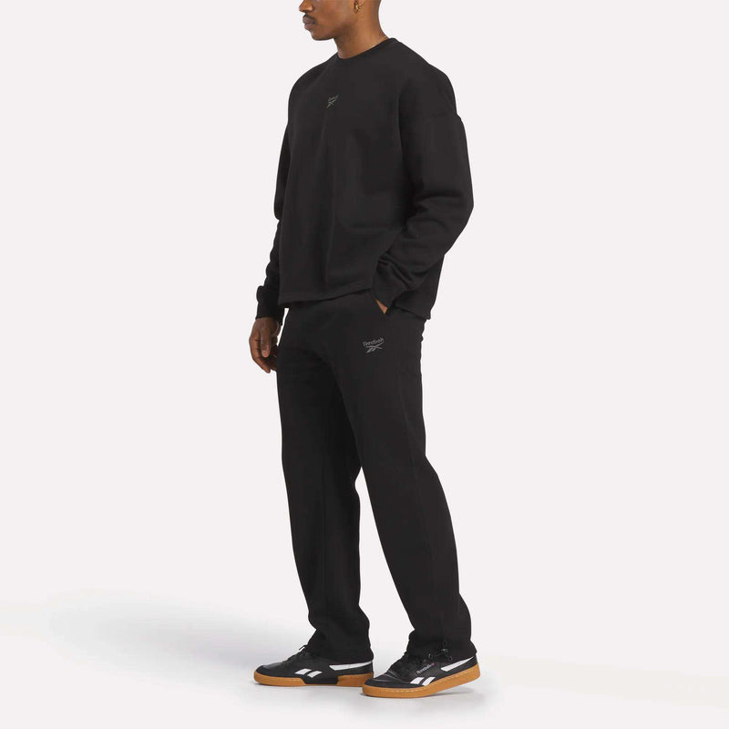 A person stands against a white background wearing black sneakers, a black sweatshirt, and Reebok Men's Wardrobe Essentials Pants for a casual and relaxed look.