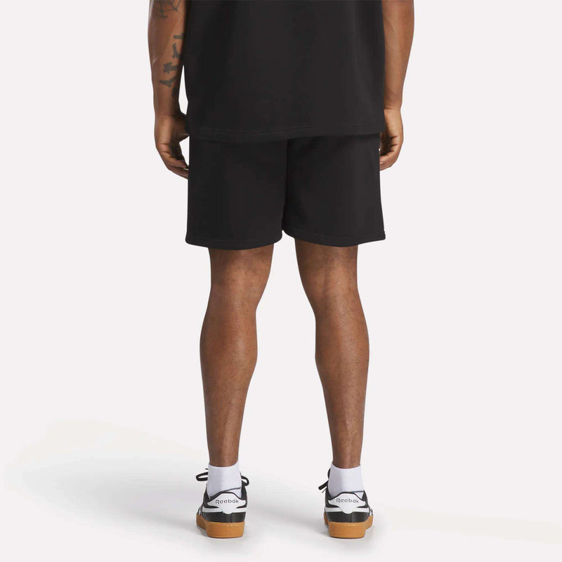 A person stands with their back to the camera, showing only their lower back, legs, and feet. They're wearing Reebok Men's Wardrobe Essentials Shorts with a relaxed fit, styled with a black tee, white socks, and black-and-white sneakers on a white background.