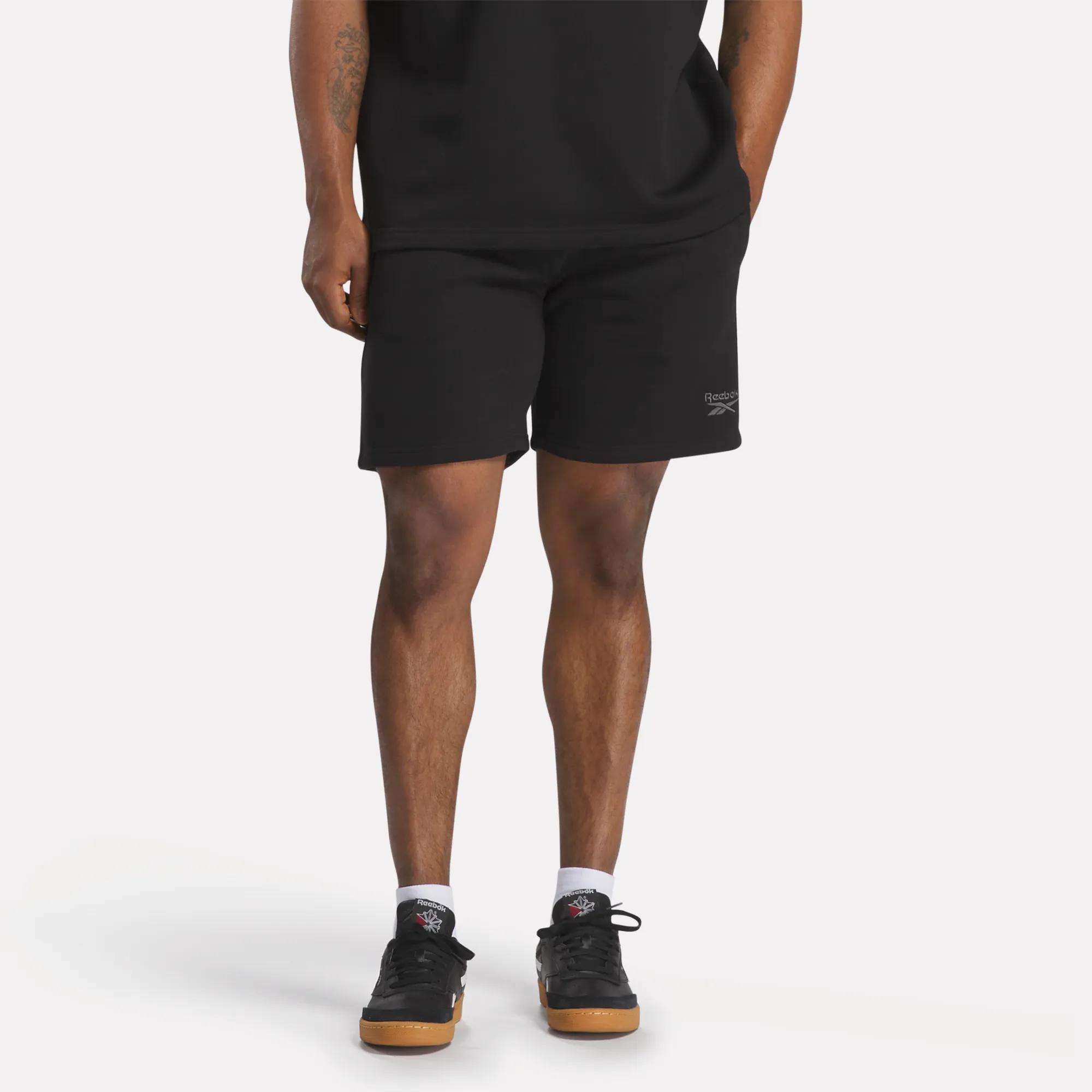A person in a black t-shirt, Reebok Men's Wardrobe Essentials Shorts (relaxed fit), white socks, and black sneakers stands against a plain light background. Only their body from the shoulders down is visible.