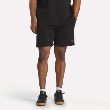 A person in a black t-shirt, Reebok Men's Wardrobe Essentials Shorts (relaxed fit), white socks, and black sneakers stands against a plain light background. Only their body from the shoulders down is visible.