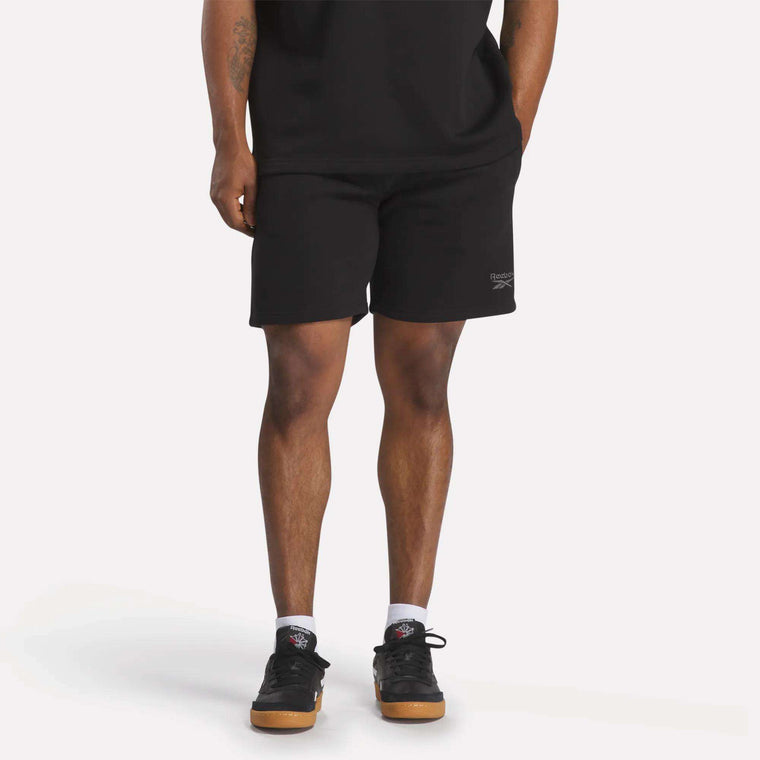 A person in a black t-shirt, Reebok Men's Wardrobe Essentials Shorts (relaxed fit), white socks, and black sneakers stands against a plain light background. Only their body from the shoulders down is visible.