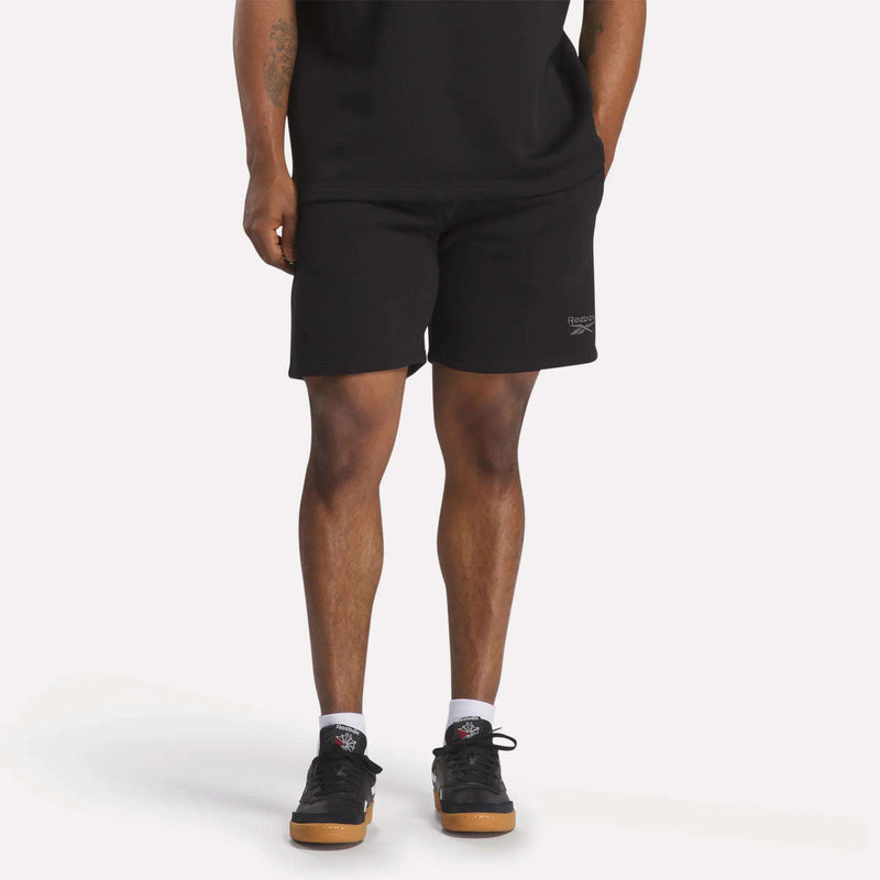 A person in a black t-shirt, Reebok Men's Wardrobe Essentials Shorts (relaxed fit), white socks, and black sneakers stands against a plain light background. Only their body from the shoulders down is visible.