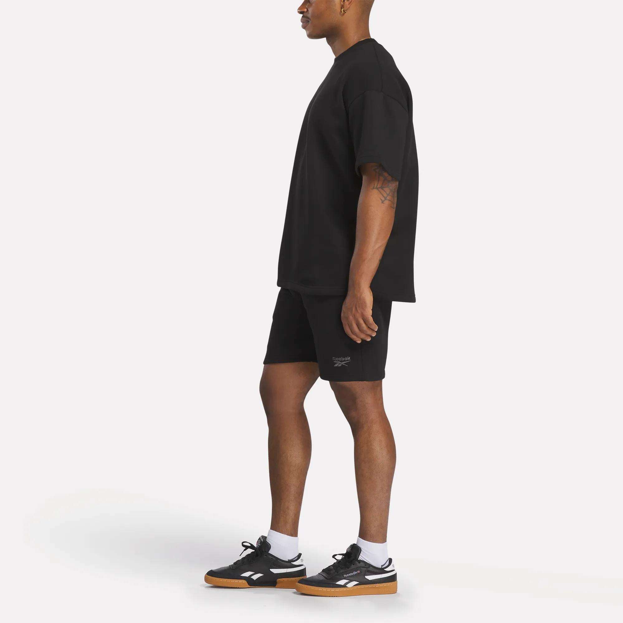 A man stands sideways in a loose black t-shirt, Reebok Men's Wardrobe Essentials Shorts, white socks, and black sneakers with gum soles against a plain white background.