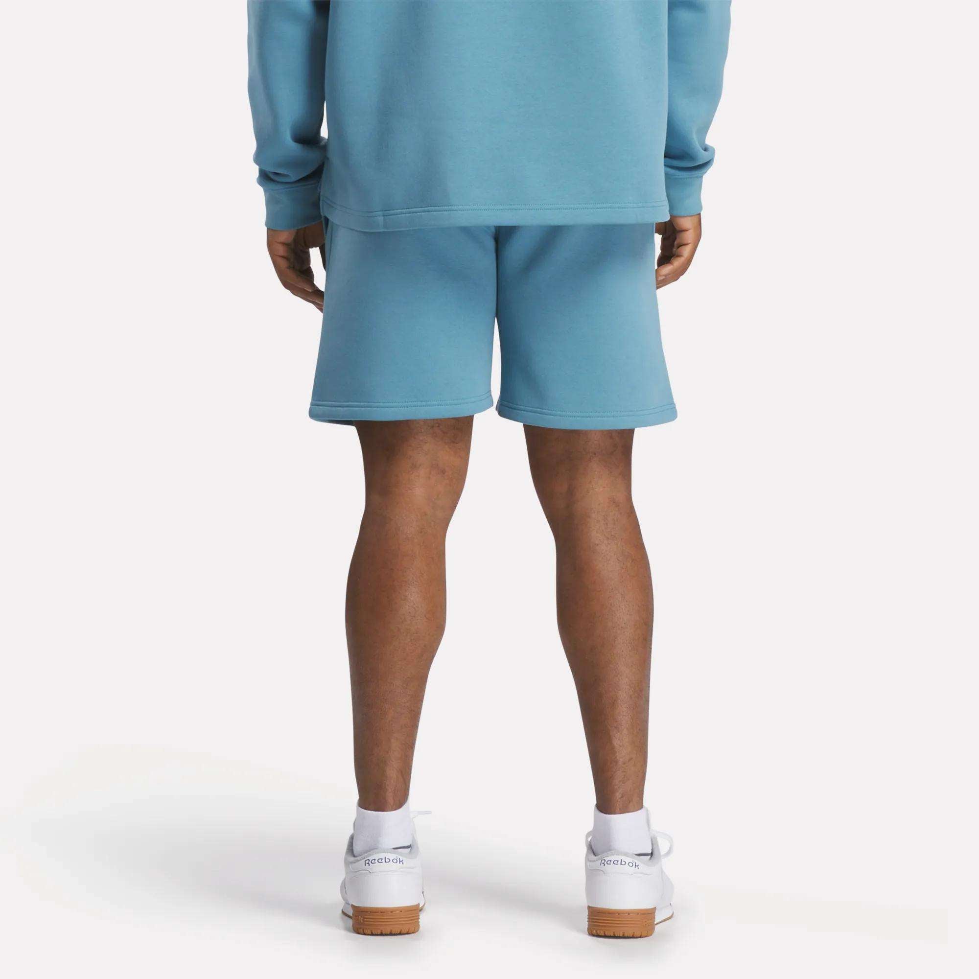 A person stands facing away, wearing a relaxed fit light blue sweatshirt, Reebok Men's Wardrobe Essentials Shorts in matching blue, and white sneakers against a plain white background.