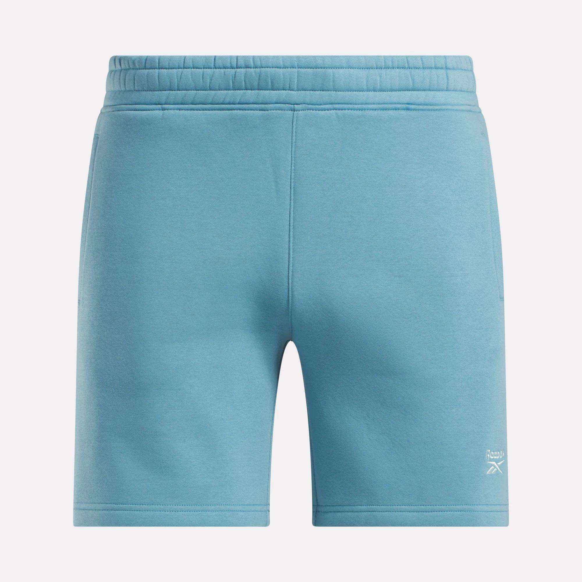 Reebok Men's Wardrobe Essentials Shorts in light blue, featuring a relaxed fit, elastic waistband, subtle stitching, and a small white logo on the lower left leg. Displayed from the front against a plain white background.