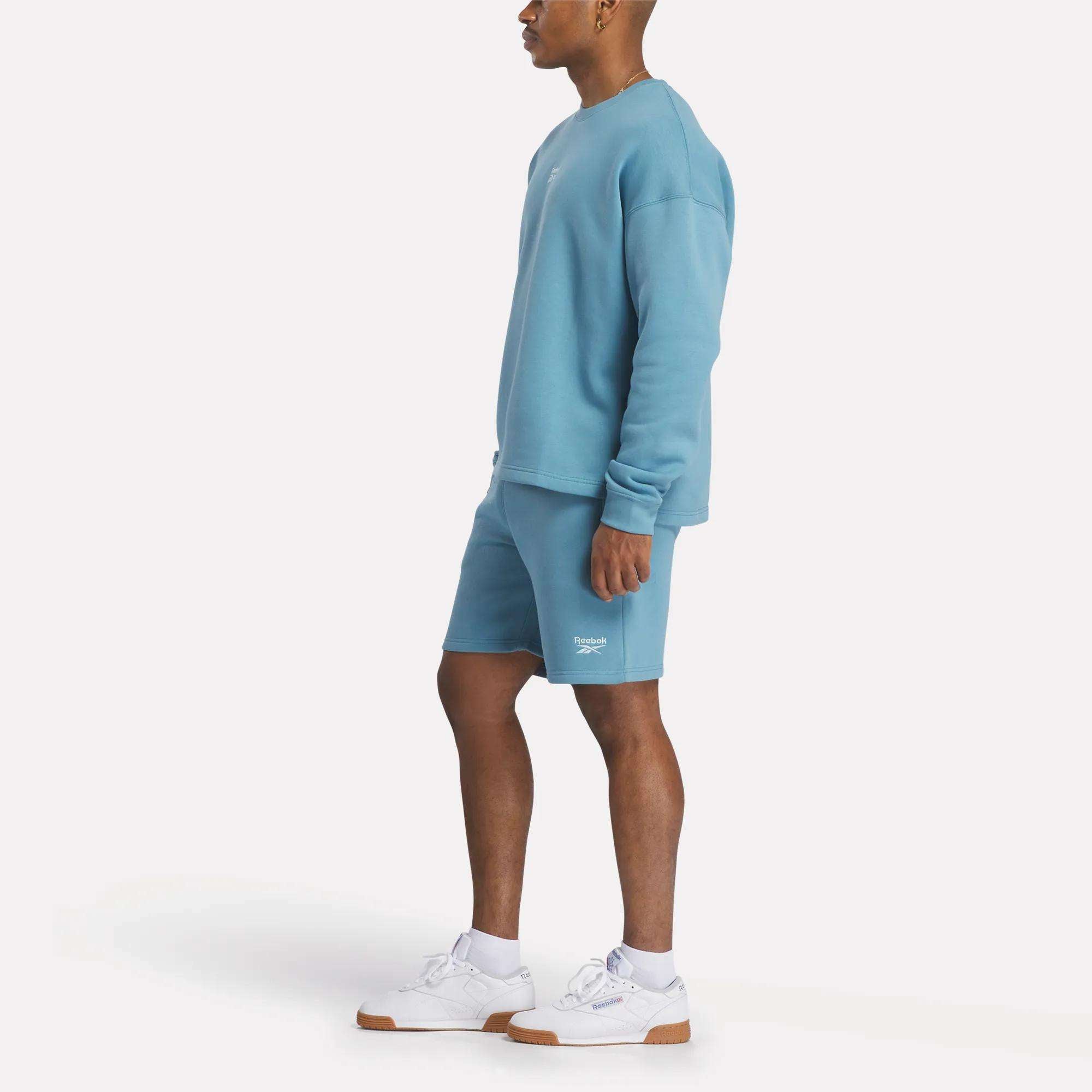 A man stands sideways against a plain white background wearing a light blue sweatshirt, white sneakers, and Reebok Men's Wardrobe Essentials Shorts.