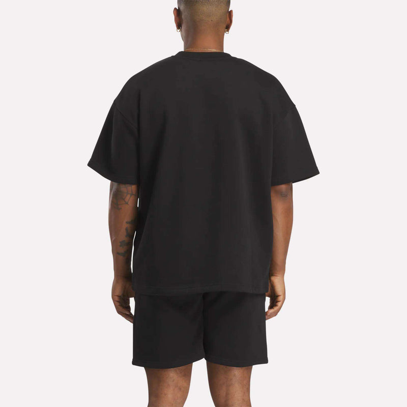 A person stands with their back to the camera, wearing a relaxed fit Men's Wardrobe Essentials T-Shirt by Reebok in black. The plain light background emphasizes the outfit, and their arms hang naturally at their sides.