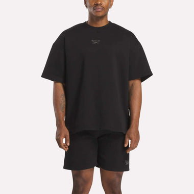 A man faces forward in a relaxed fit black Reebok Men's Wardrobe Essentials T-Shirt and black shorts, both featuring small white logos on the chest and left leg. The background is plain white.