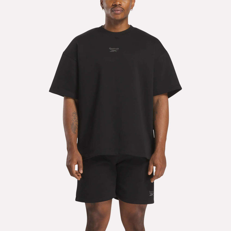 A man faces forward in a relaxed fit black Reebok Men's Wardrobe Essentials T-Shirt and black shorts, both featuring small white logos on the chest and left leg. The background is plain white.
