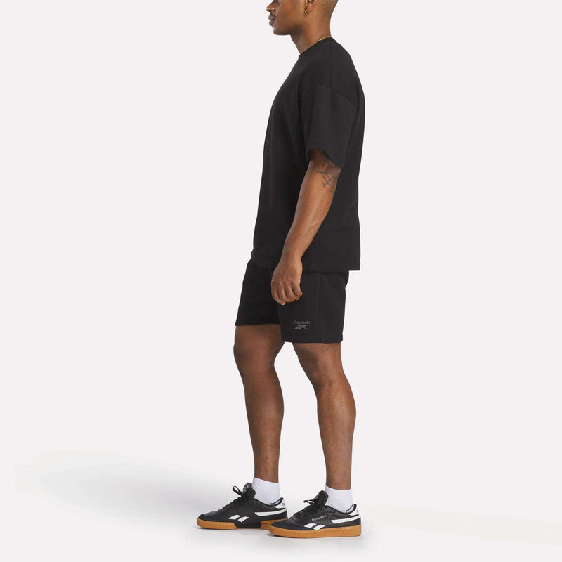 A man stands in profile against a plain white background, wearing a relaxed fit Reebok Men's Wardrobe Essentials T-Shirt, black shorts, white socks, and black sneakers with gum soles.