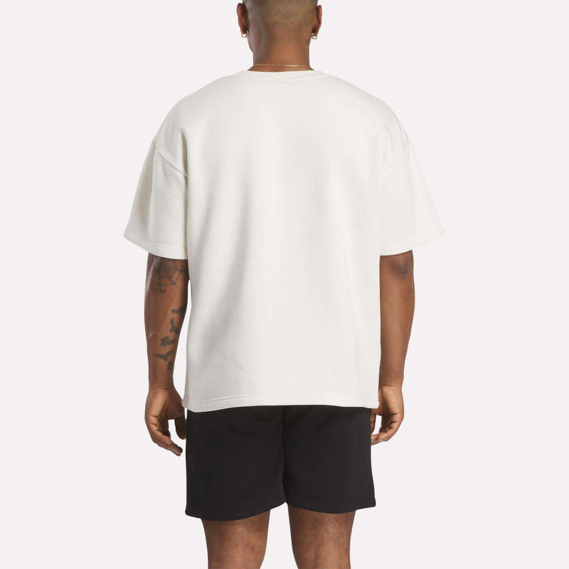 A person with short hair is seen from the back, wearing a white Reebok Men's Wardrobe Essentials T-Shirt and black shorts. Their arms hang relaxed by their sides, showing tattoos on the left arm. The background is plain and light-colored.