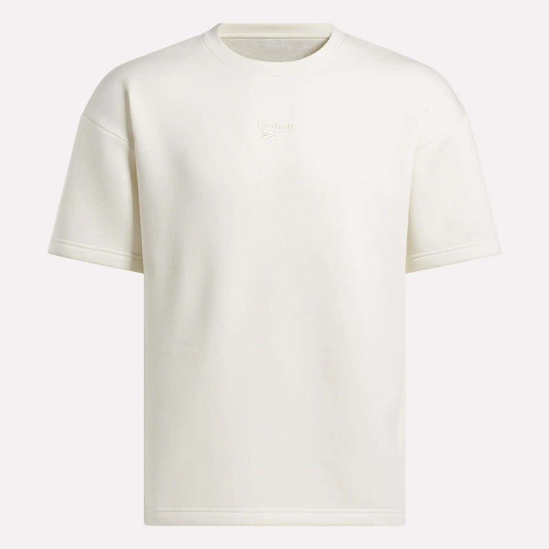 The Reebok Men's Wardrobe Essentials T-Shirt is an off-white, relaxed-fit crewneck with short sleeves. It features subtle text and an embroidered Reebok logo on the chest for a minimal look.