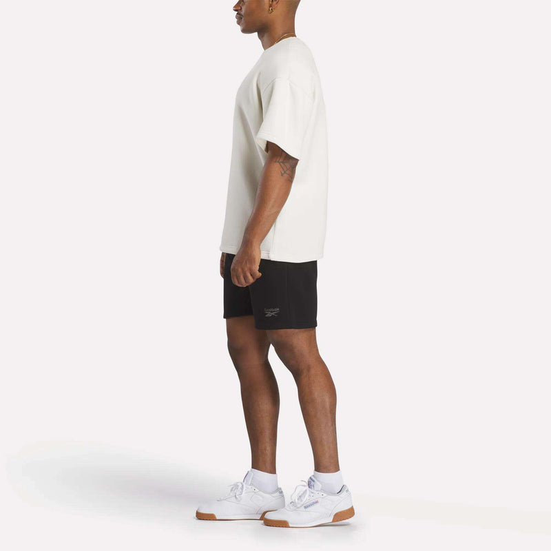 A man stands in profile against a white background, wearing a relaxed fit Reebok Men's Wardrobe Essentials T-Shirt in white, black shorts, and white athletic sneakers with gum soles.