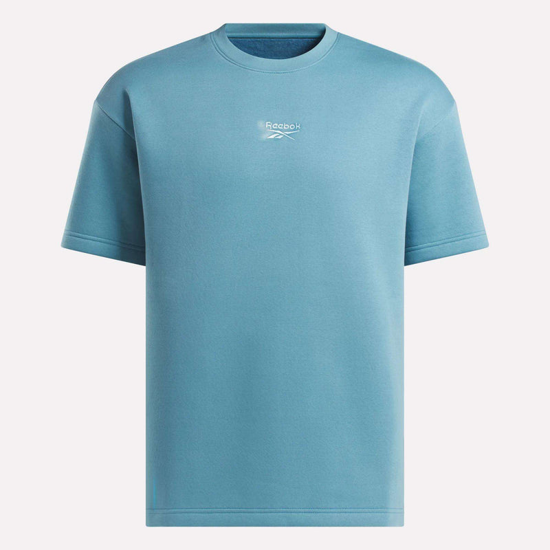 The Men's Wardrobe Essentials T-Shirt by Reebok is a plain light blue tee with a relaxed fit, short sleeves, and crew neck. It features a small white Reebok logo and text centered on the chest, shown against a white background.