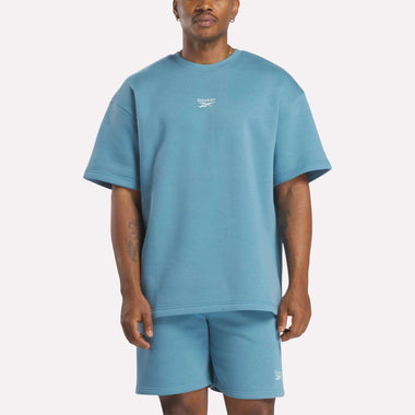 A man wears a relaxed fit, light blue Reebok Men's Wardrobe Essentials T-Shirt and matching shorts, standing facing forward against a plain white background.