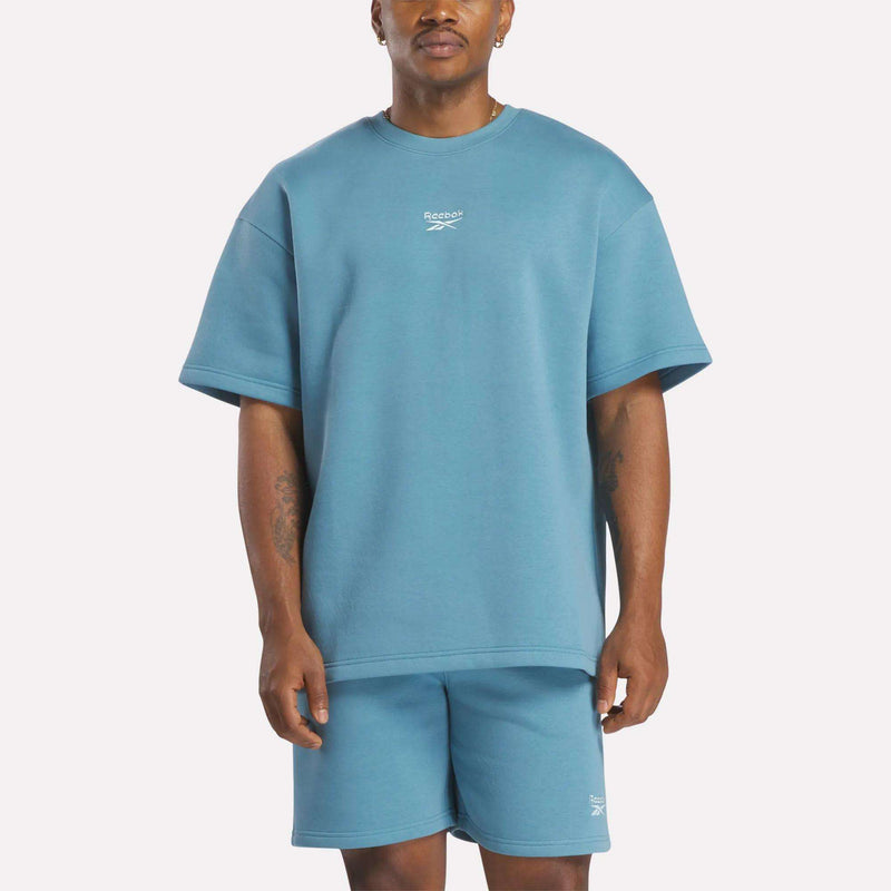 A man wears a relaxed fit, light blue Reebok Men's Wardrobe Essentials T-Shirt and matching shorts, standing facing forward against a plain white background.