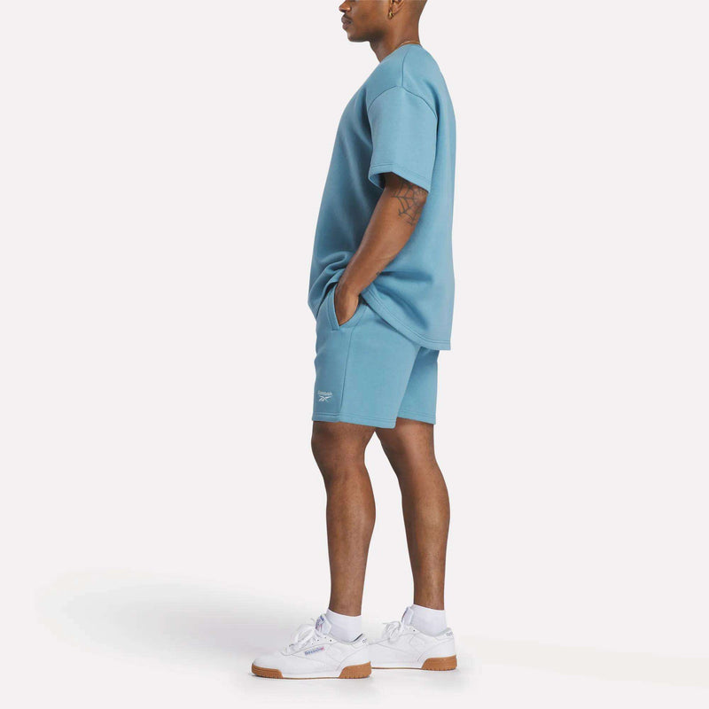 A man stands in profile against a white background wearing the relaxed fit Men's Wardrobe Essentials T-Shirt by Reebok in light blue, paired with matching shorts and white sneakers, hands in his pockets.