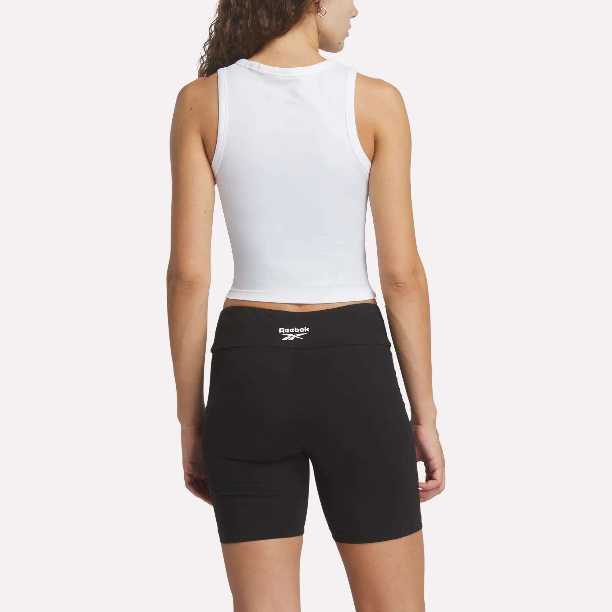 A woman faces away, wearing the Reebok Women's Rib Tank Top in white with black cycling shorts against a plain, light background.