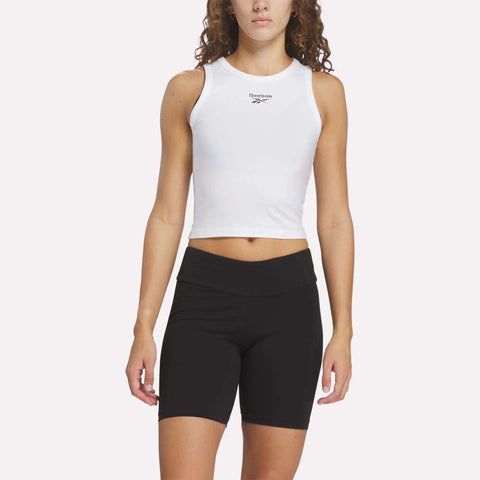 A person with long curly hair wears a sleeveless white Reebok Women's Rib Tank Top and high-waisted black bike shorts, standing against a plain light background.