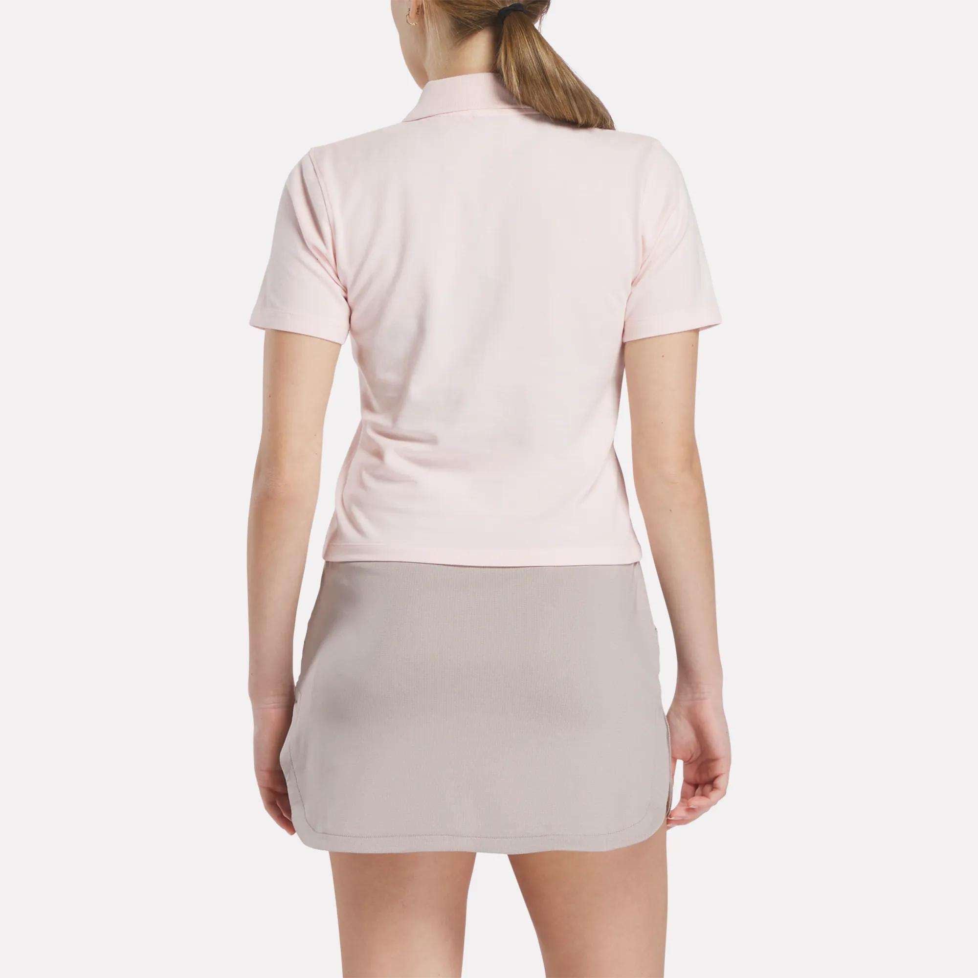 A woman with a ponytail wears a Reebok Women's Fitted Polo Shirt in light pink stretchy cotton and a light gray skirt, standing with her back to the camera against a plain white background.
