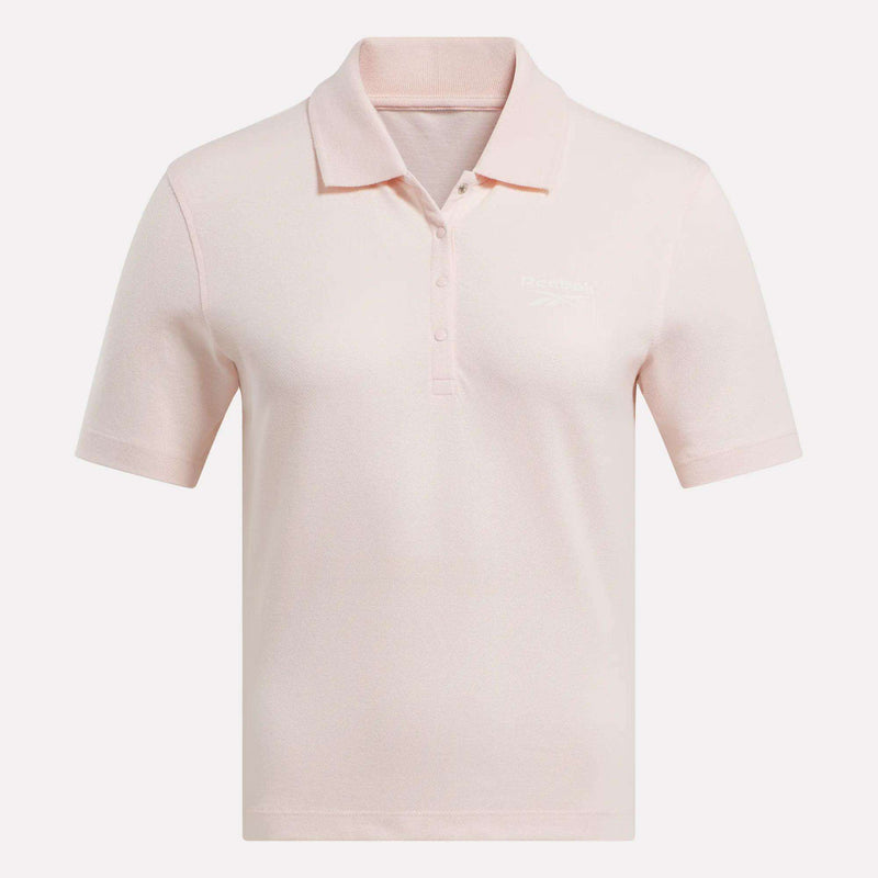 The Women's Fitted Polo Shirt by Reebok is a light pink, stretchy cotton top with short sleeves, a three-button collar, and a subtle embroidered logo on the left chest, shown on a plain white background.