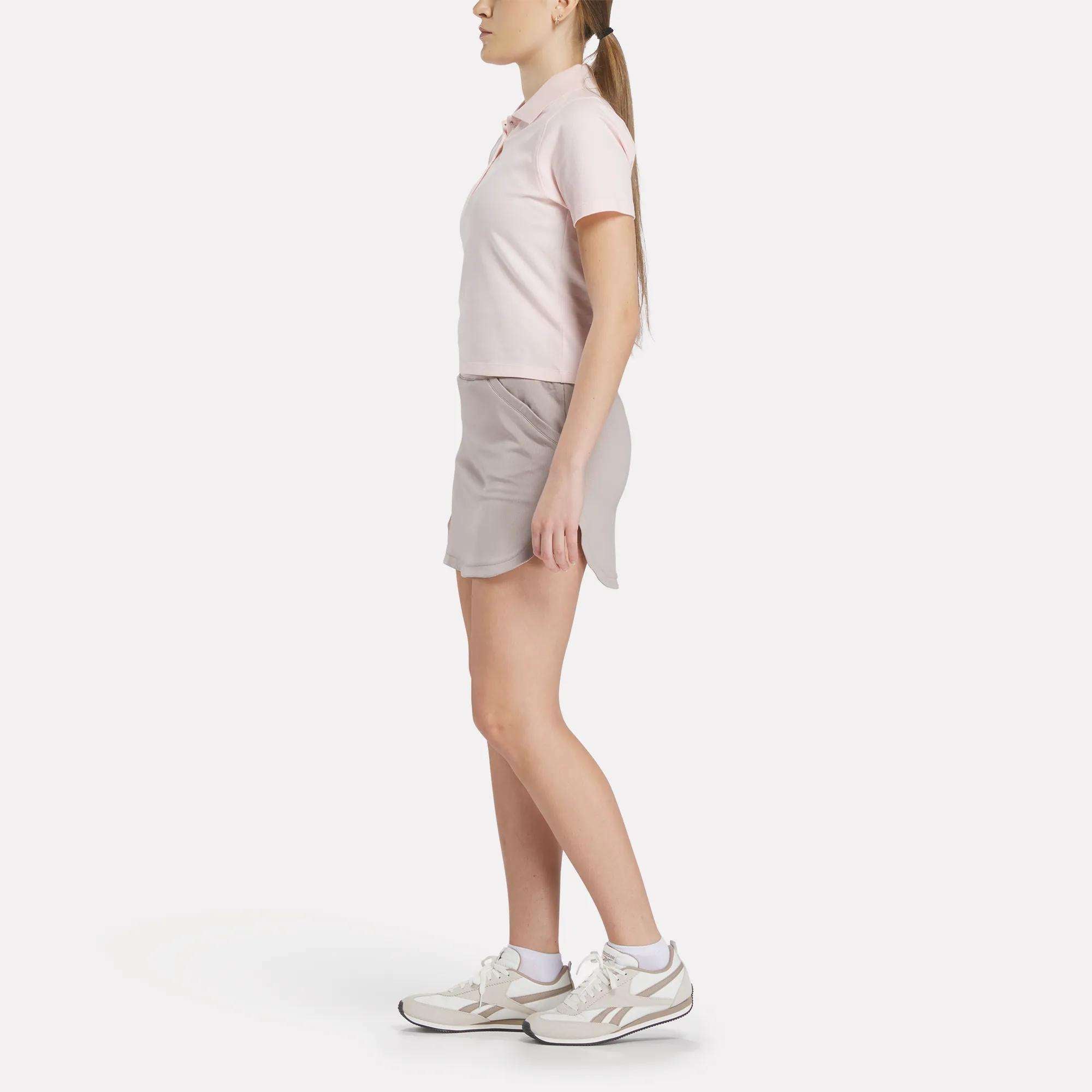 A woman in profile stands against a white background, wearing a light pink Reebok Women's Fitted Polo Shirt, a light gray skirt, and white sneakers; her hair is tied back in a ponytail.