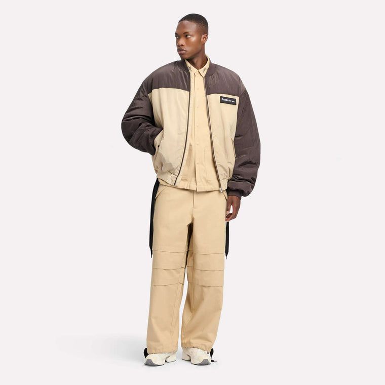 A man wears the Reebok Men's LTD Bomber Jacket in color-blocked brown and beige over a relaxed beige utilitarian shirt and wide-leg pants with white sneakers, set against a plain light background.