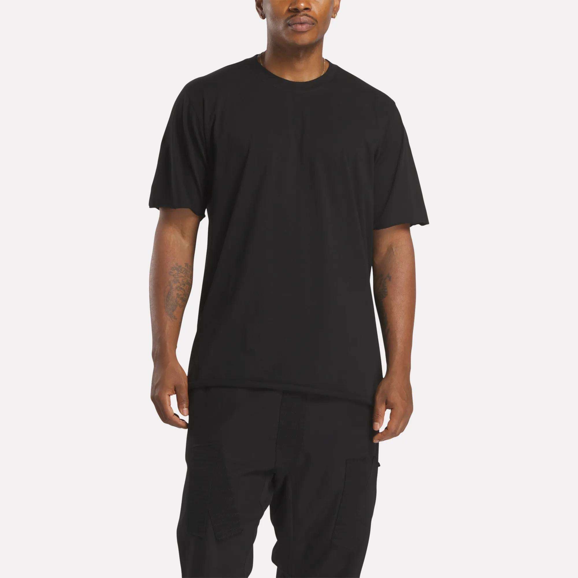 A person stands against a plain background in a loose black Reebok x 11 By Boris Bidjan Saberi LTD Constructed Tee and black pants, with arms relaxed at their sides, capturing a minimalist streetwear vibe.