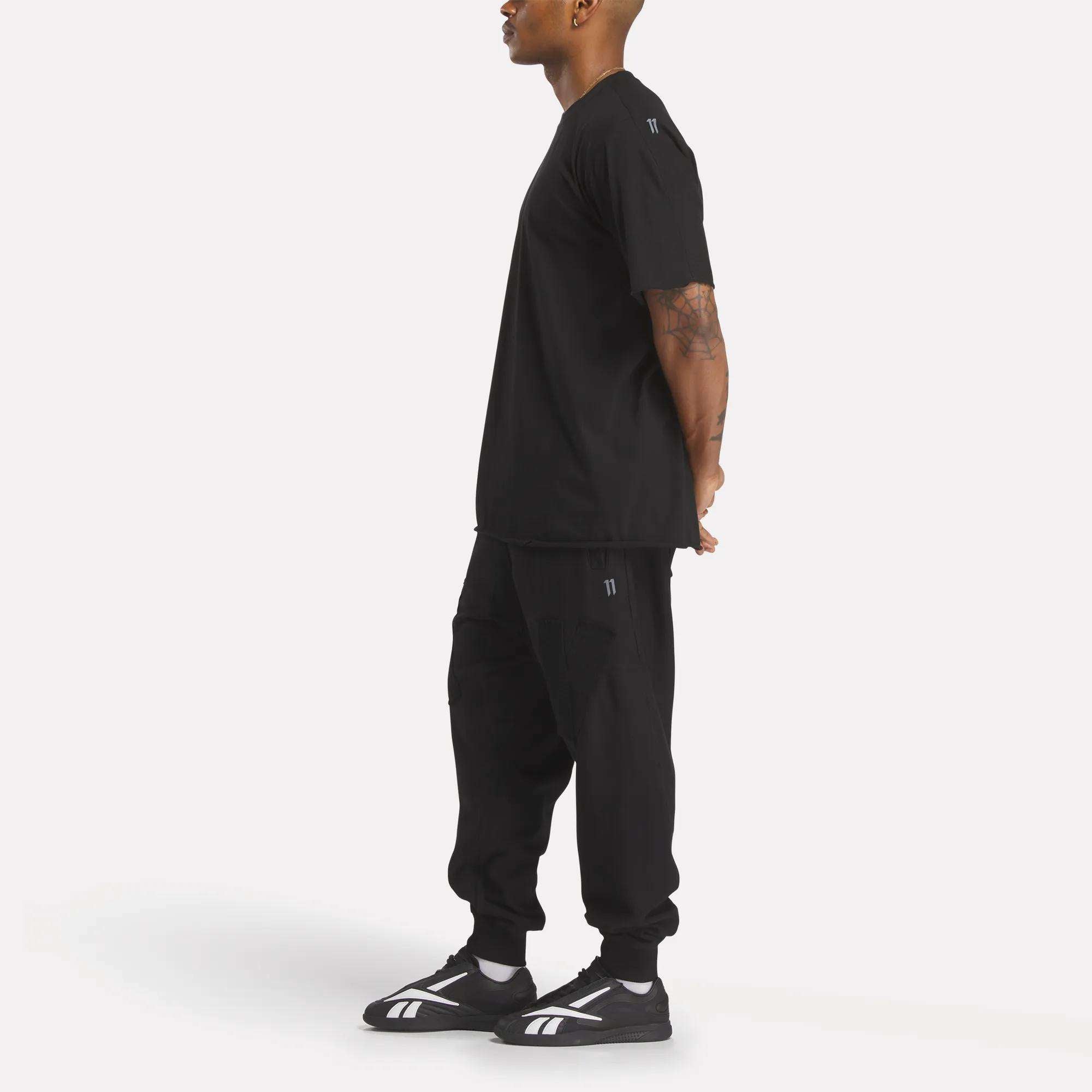 A person stands in profile with hands behind their back, wearing the Reebok x 11 By Boris Bidjan Saberi LTD Constructed Tee, paired with black joggers and black athletic shoes featuring white stripes, against a plain white background.