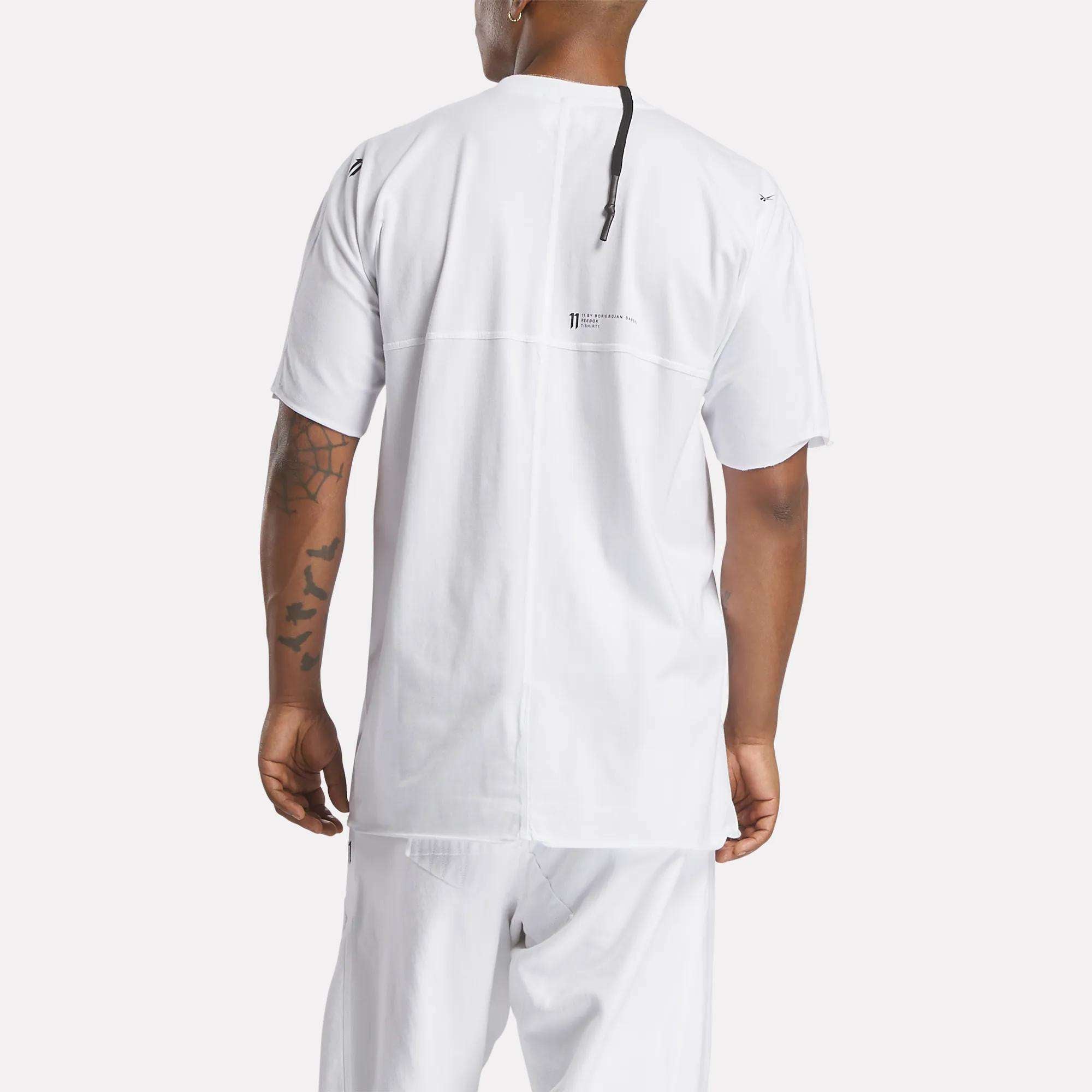 A person is seen from the back wearing the Reebok x 11 By Boris Bidjan Saberi LTD Constructed Tee in white with relaxed white pants. The tee has a small upper-center logo and text, plus visible tattoos on their left arm—streetwear essentials.
