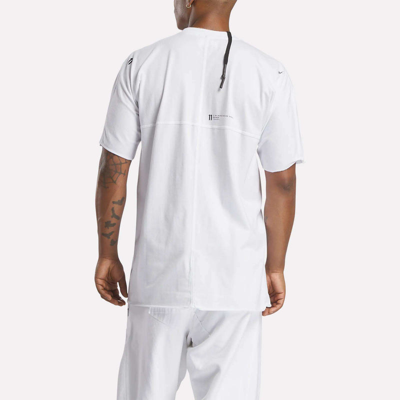 A person is seen from the back wearing the Reebok x 11 By Boris Bidjan Saberi LTD Constructed Tee in white with relaxed white pants. The tee has a small upper-center logo and text, plus visible tattoos on their left arm—streetwear essentials.