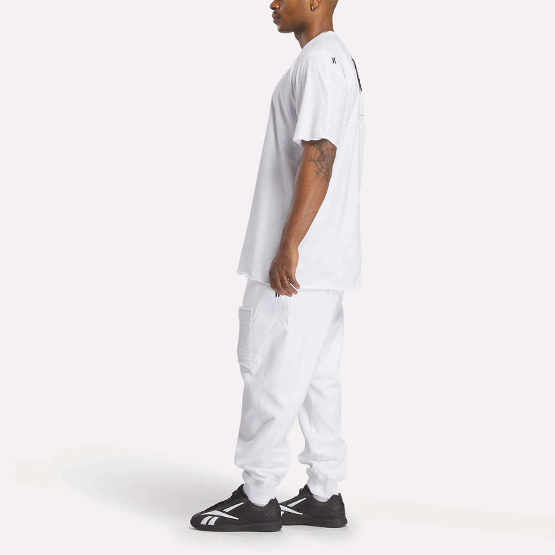 A man stands sideways in a loose-fitting Reebok x 11 By Boris Bidjan Saberi LTD Constructed Tee, white cargo pants, and black-and-white sneakers, exuding modern streetwear style against a plain white background.