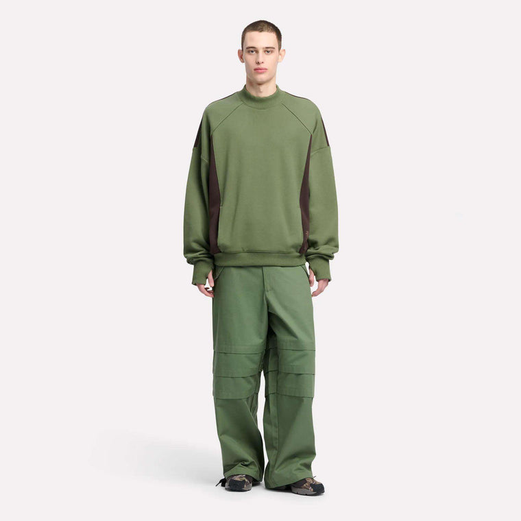 A person stands facing forward in a Reebok Men's Uniform LTD Colorblock Sweatshirt and loose green pants with seam detailing, paired with black sandals, all set against a plain white background.