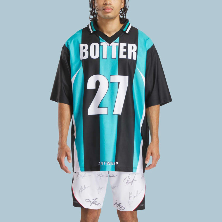 A person wears the Reebok x Botter LTD Reverse Soccer Men's Tee in teal and black stripes with BOTTER and 27, paired with signed white shorts. This standout Reebok look pops against a light blue backdrop.