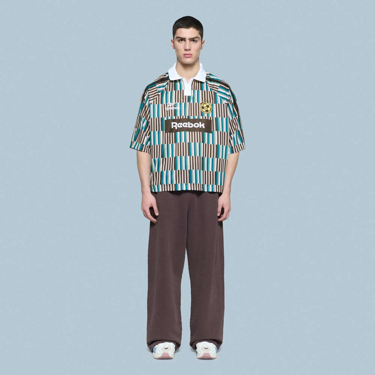 A young man faces forward, wearing a blue, white, and brown Reebok Men's Soccer LTD T-Shirt 22 with wide brown pants and white sneakers, set against a plain light blue background.