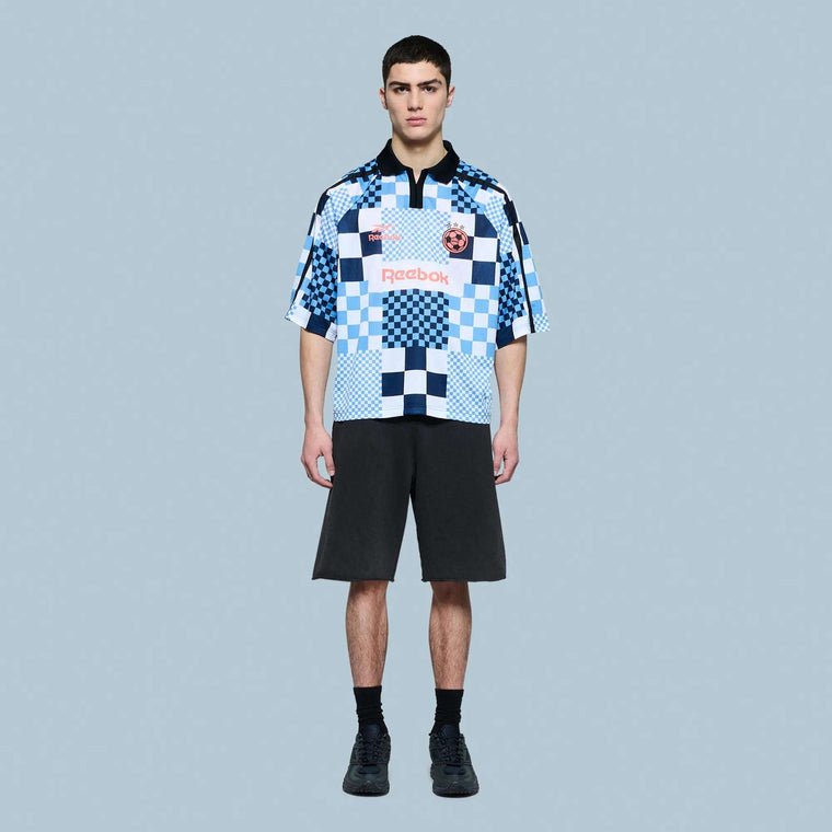 A young man stands facing forward against a plain light blue background, wearing a Reebok Men's Soccer LTD T-Shirt 22 with black shorts and black shoes.