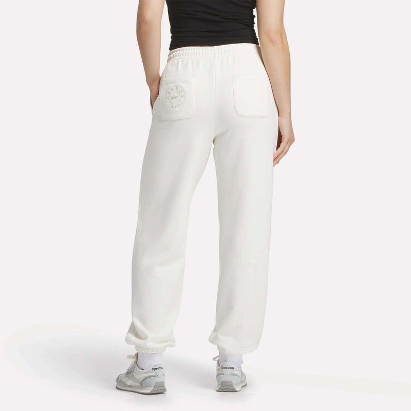 A person stands with their back to the camera against a white background, wearing a black top, white Reebok Women's Lawn Club Jogger Pants with back pockets, white socks, and gray sneakers.