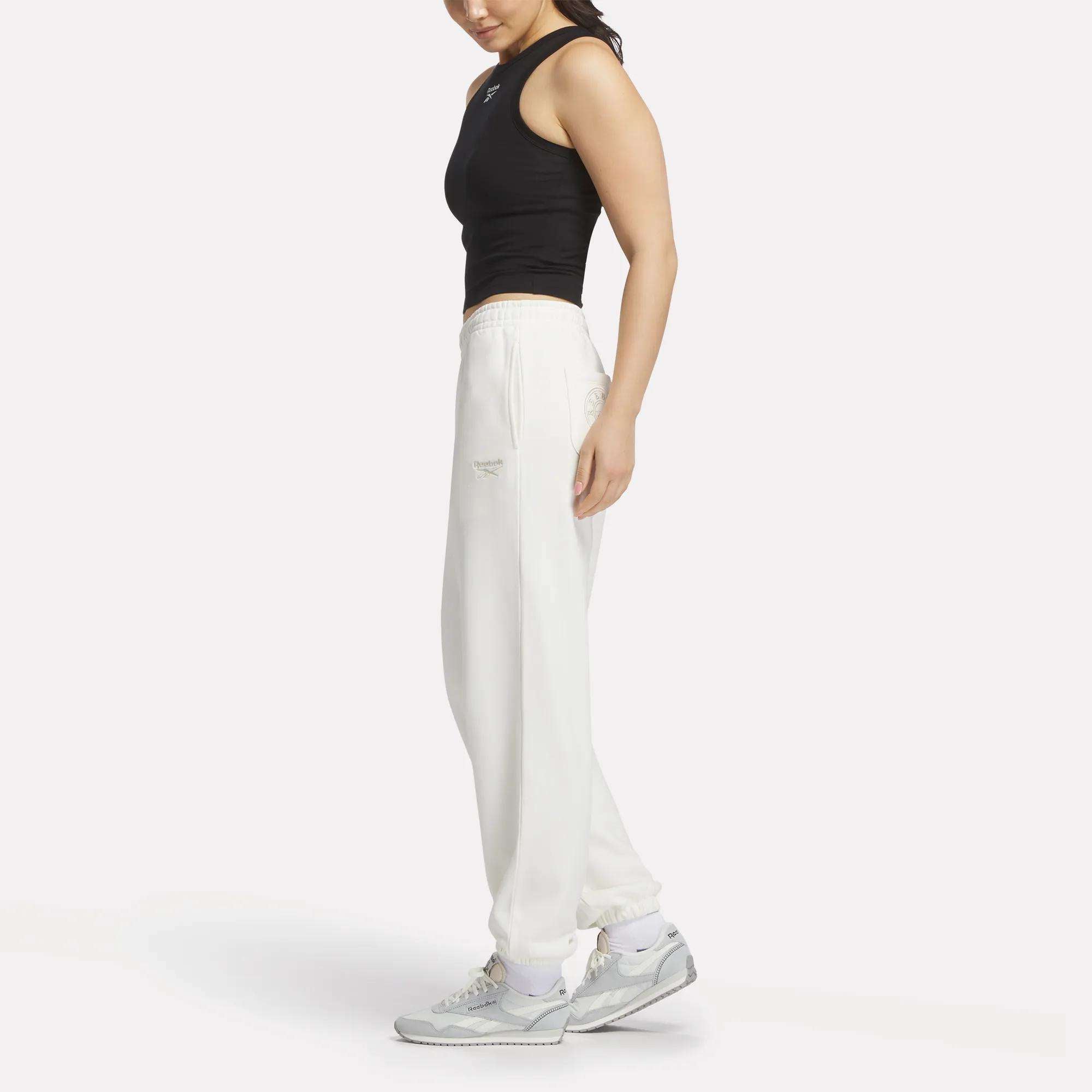 A person in a black sleeveless top, Reebok Women's Lawn Club Jogger Pants in white, and gray sneakers stands sideways against a plain white background.