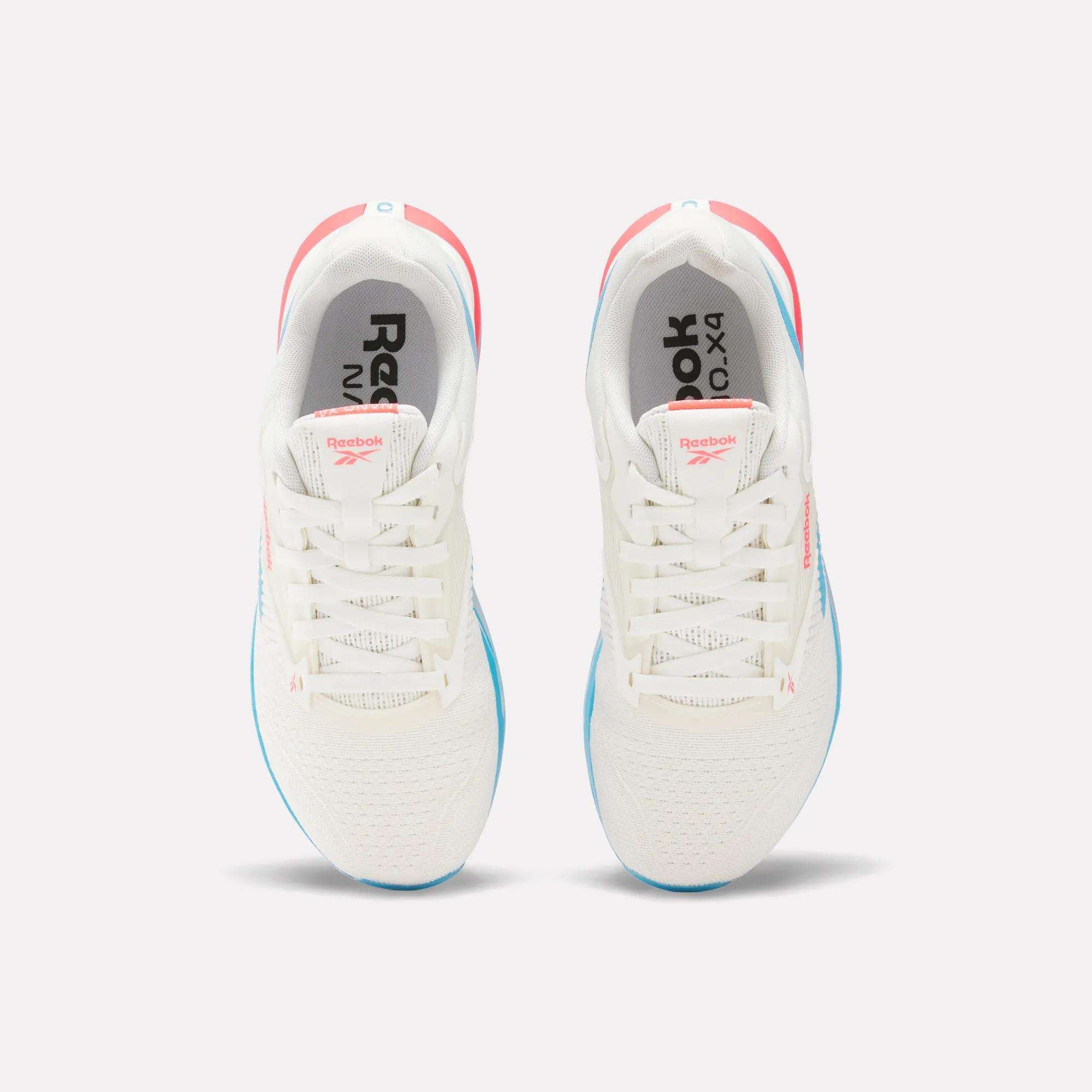 Top view of a pair of Women’s Nano X4 Training Shoes by Reebok in white with blue soles and pink accents, placed side by side. The breathable shoes show the Reebok logo on the insoles and tongues.