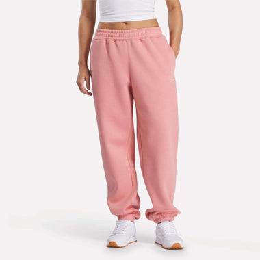 A person models Reebok Women's Wardrobe Essentials Sweat Pants in pink, paired with a white cropped top and white sneakers, standing against a plain background with hands in pockets and face not visible.