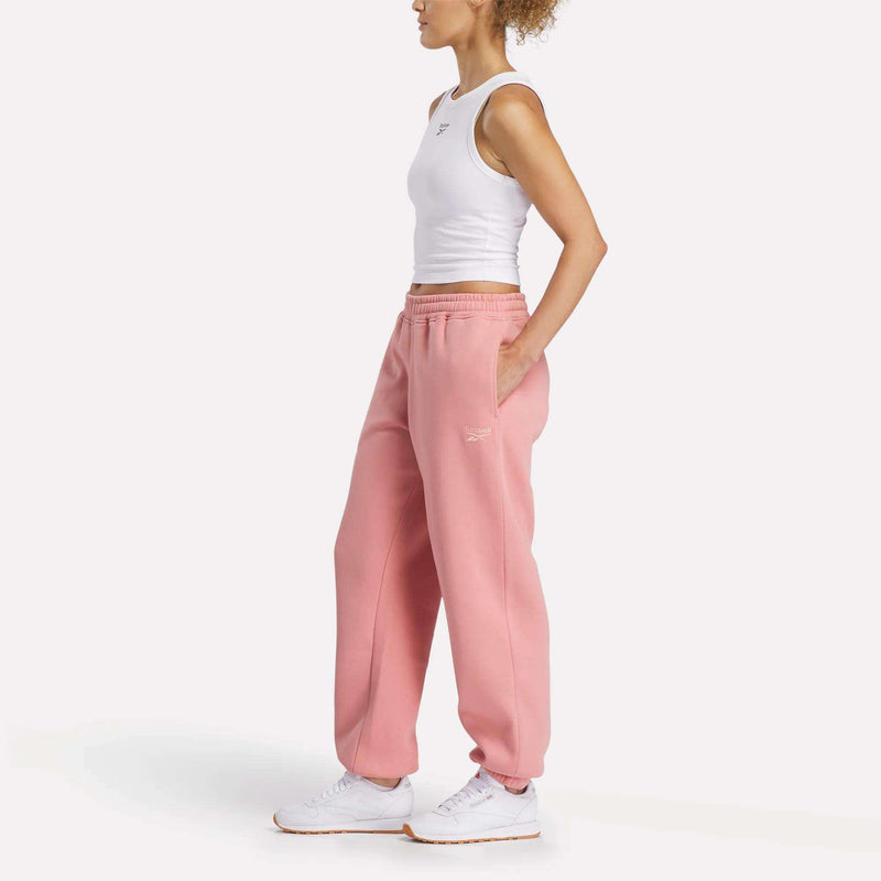 A woman stands in profile against a white background, wearing a white sleeveless crop top, Reebok Women's Wardrobe Essentials Sweat Pants in pink with hands in pockets, and white sneakers.