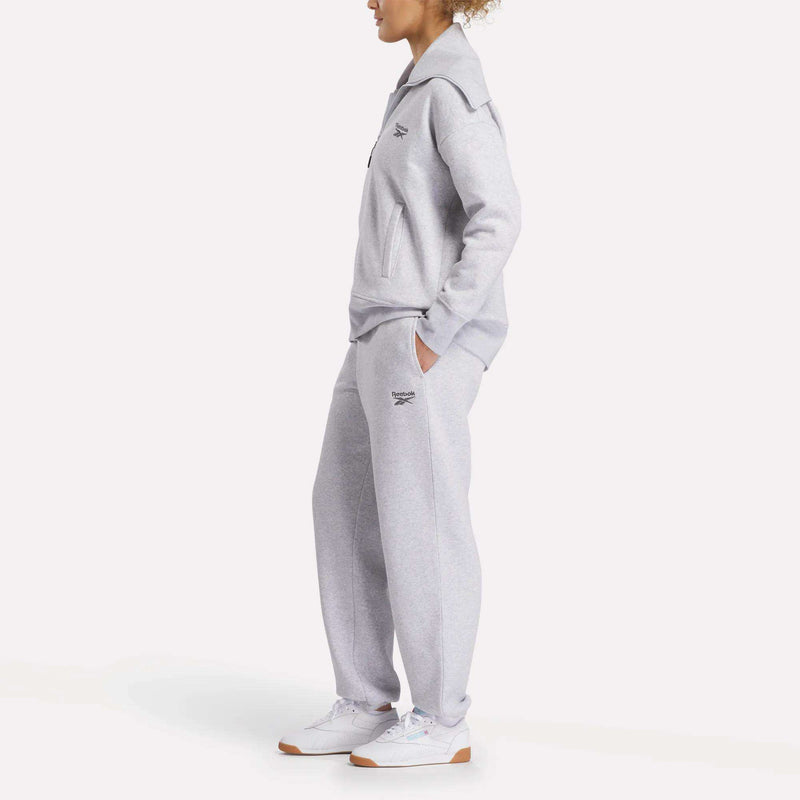 A person stands hands in pockets, wearing Reebok Women's Wardrobe Essentials Sweat Pants in light gray with a matching top and white sneakers. Small Reebok logos appear on the chest and leg as they face slightly to the side.