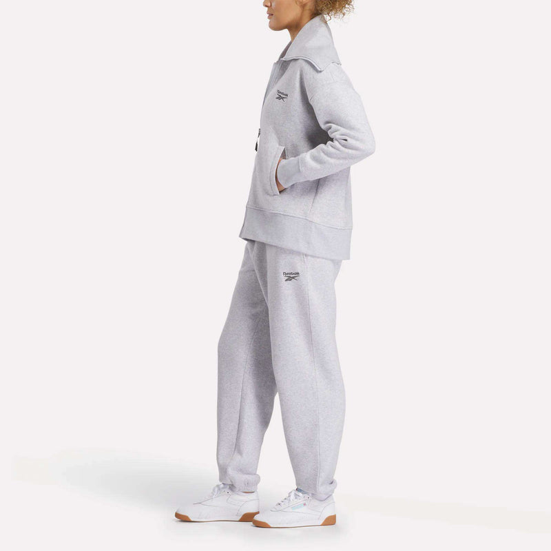 A woman stands sideways in a Reebok Women's Wardrobe Essentials Retro Zip Top, paired with matching sweatpants and white sneakers. The light gray outfit features small embroidered logos on the chest and thigh, with hands in pockets against a plain white background.