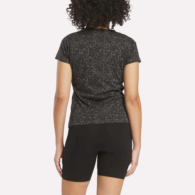 A person with curly hair is shown from the back wearing the Reebok Women's Leopard Print Baby T-Shirt and black shorts, posed against a plain white background.